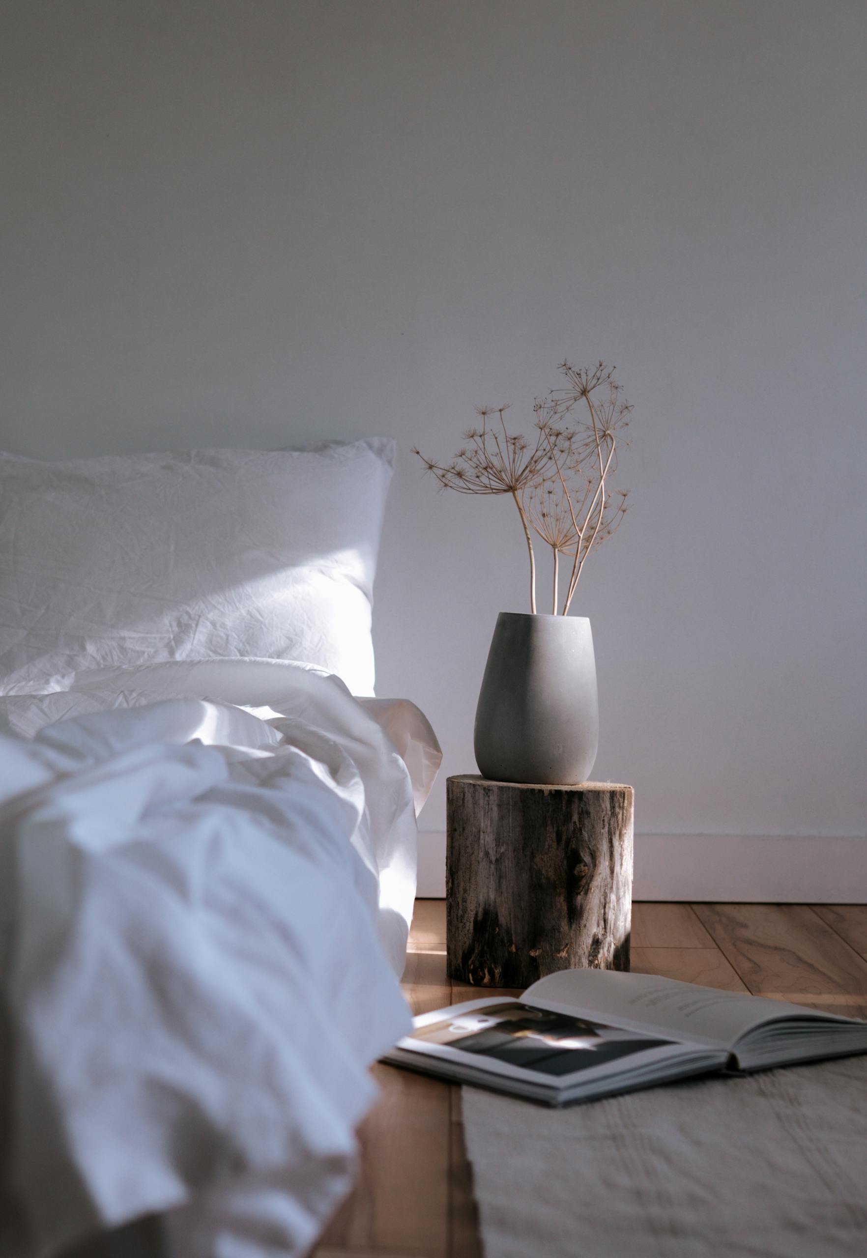 A White Bed with Linen Near the Chopped Wood with Ceramic Vase on Top