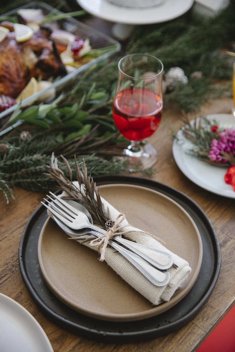 From above of served plates with cutlery and napkin tied ribbon with dry sprig near glass of wine and fir branch on festive table