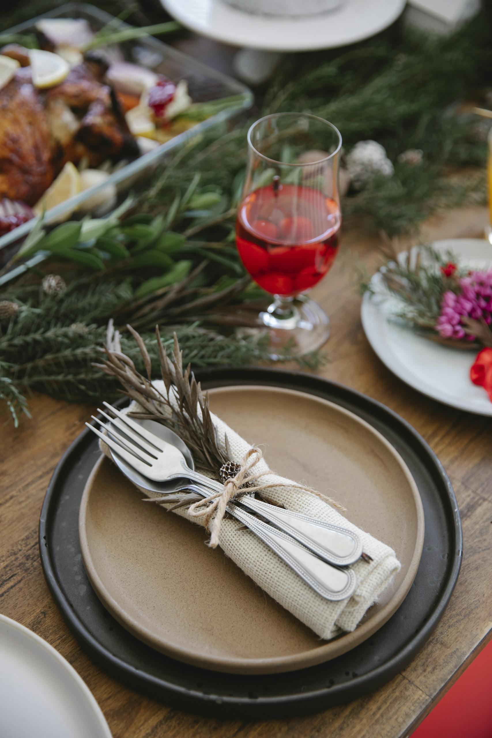 From above of served plates with cutlery and napkin tied ribbon with dry sprig near glass of wine and fir branch on festive table