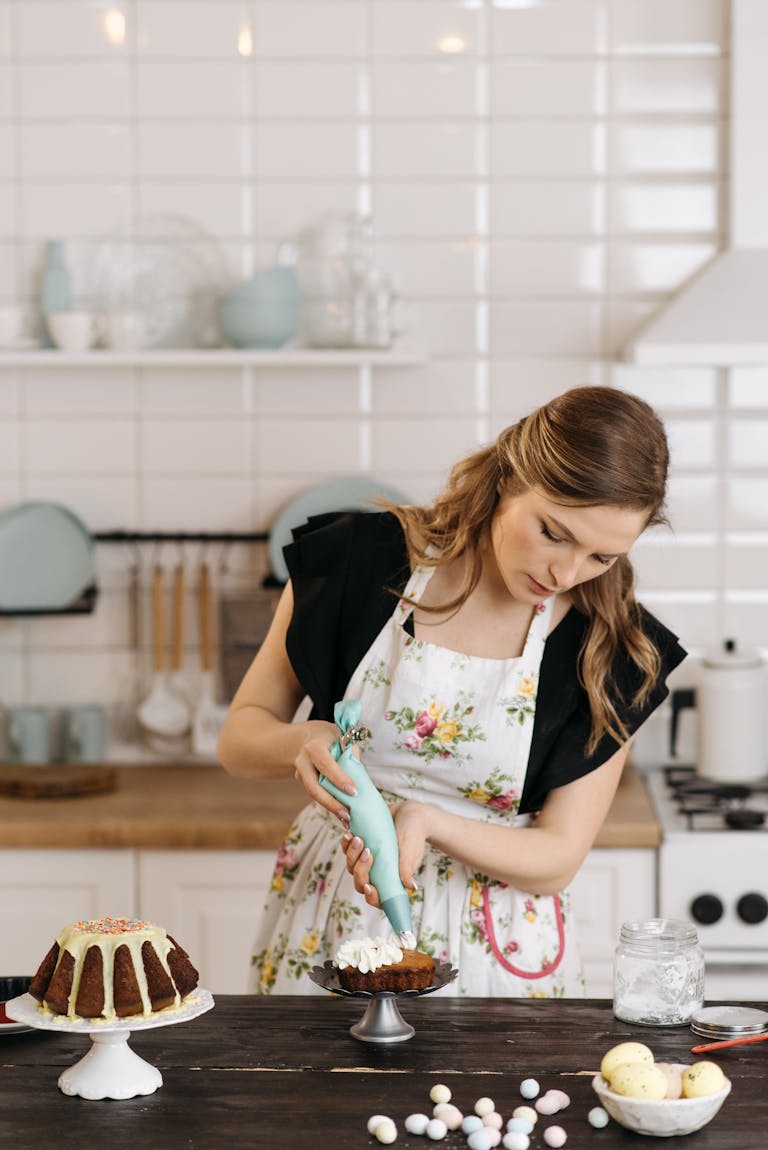 Woman in floral apron decorates cake with icing in a modern, well-lit kitchen.