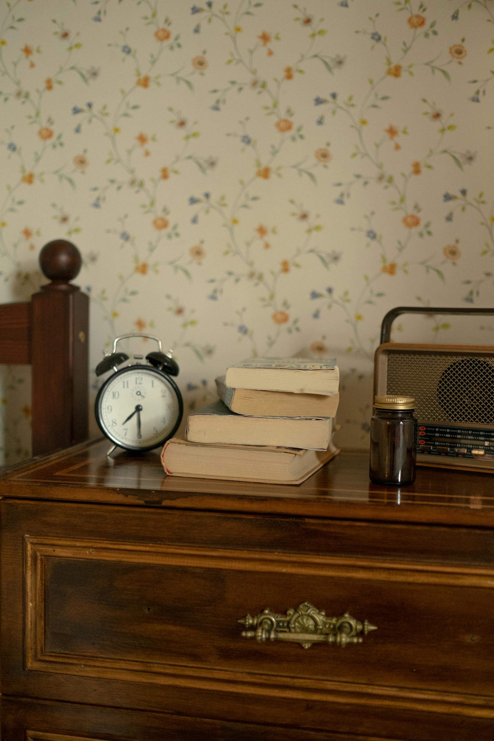 A cozy vintage bedroom setup featuring an alarm clock, books, and retro radio on a wooden chest of drawers.