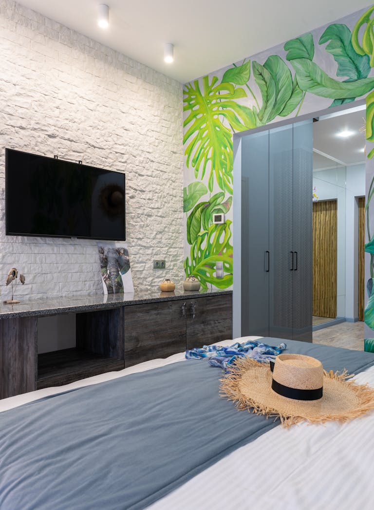 Stylish bedroom with tropical wallpaper, TV, and straw hat on bed, offering a modern yet cozy ambiance.