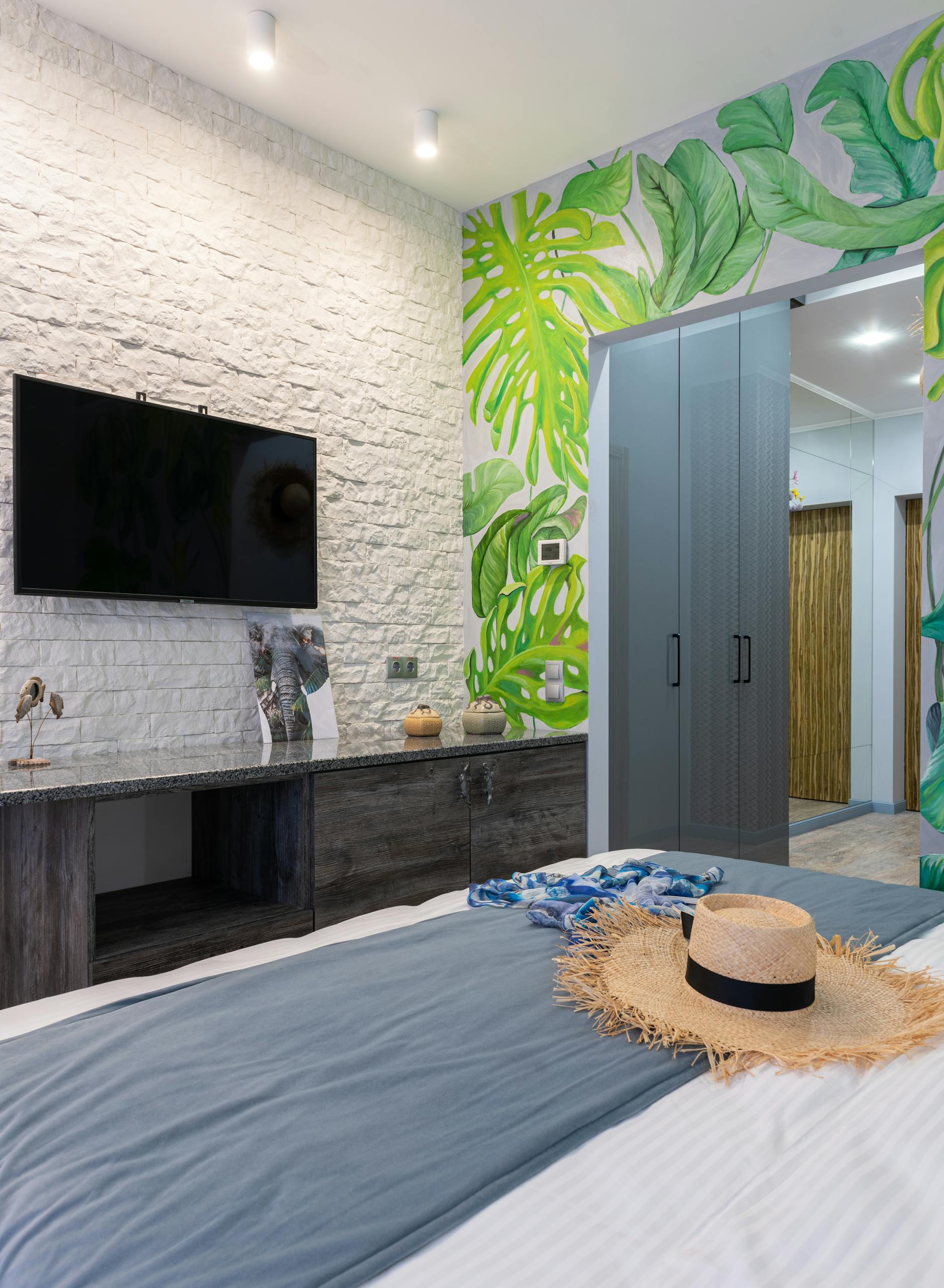 Stylish bedroom with tropical wallpaper, TV, and straw hat on bed, offering a modern yet cozy ambiance.